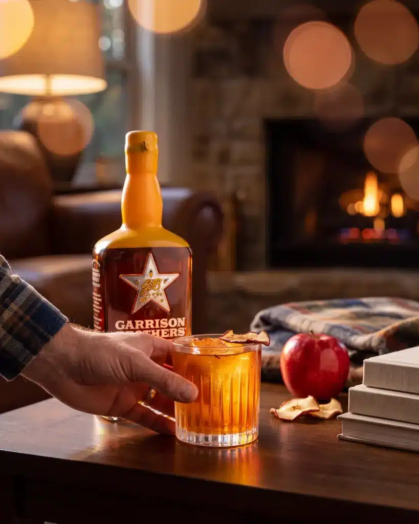 Bourbon Apple Betty bourbon cocktail by a stone fireplace with a bottle of Garrison Brothers HoneyDew Bourbon