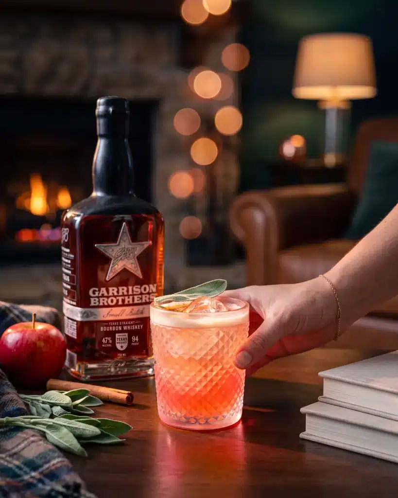 Candy Apple Ragtop bourbon cocktail by a stone fireplace with a bottle of Garrison Brothers Small Batch Bourbon