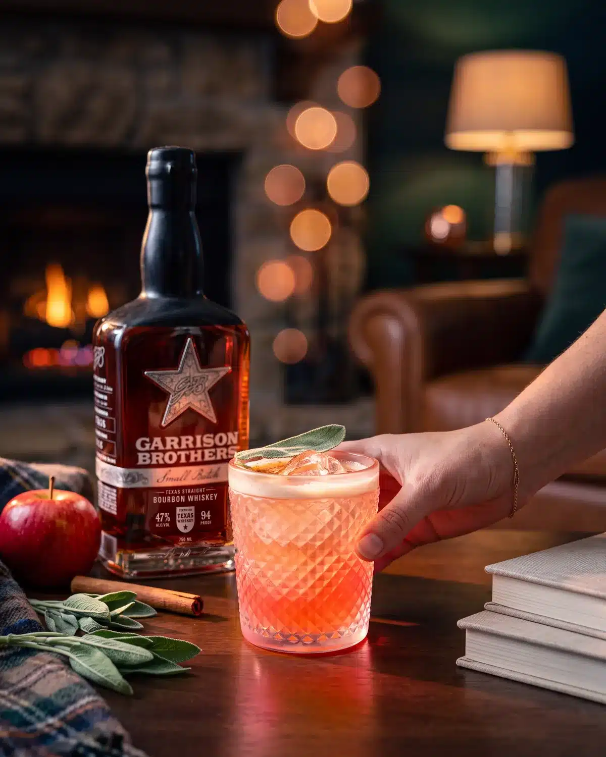 Candy Apple Ragtop bourbon cocktail by a stone fireplace with a bottle of Garrison Brothers Small Batch Bourbon