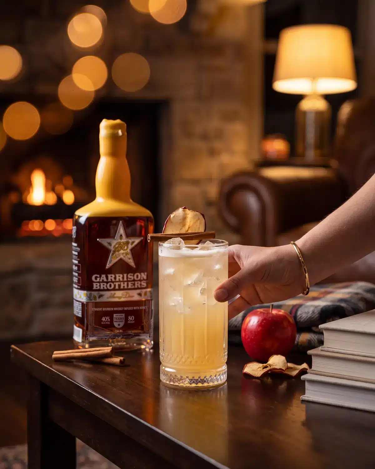 Apple Of My Hye bourbon cocktail by a stone fireplace with a bottle of Garrison Brothers HoneyDew Bourbon