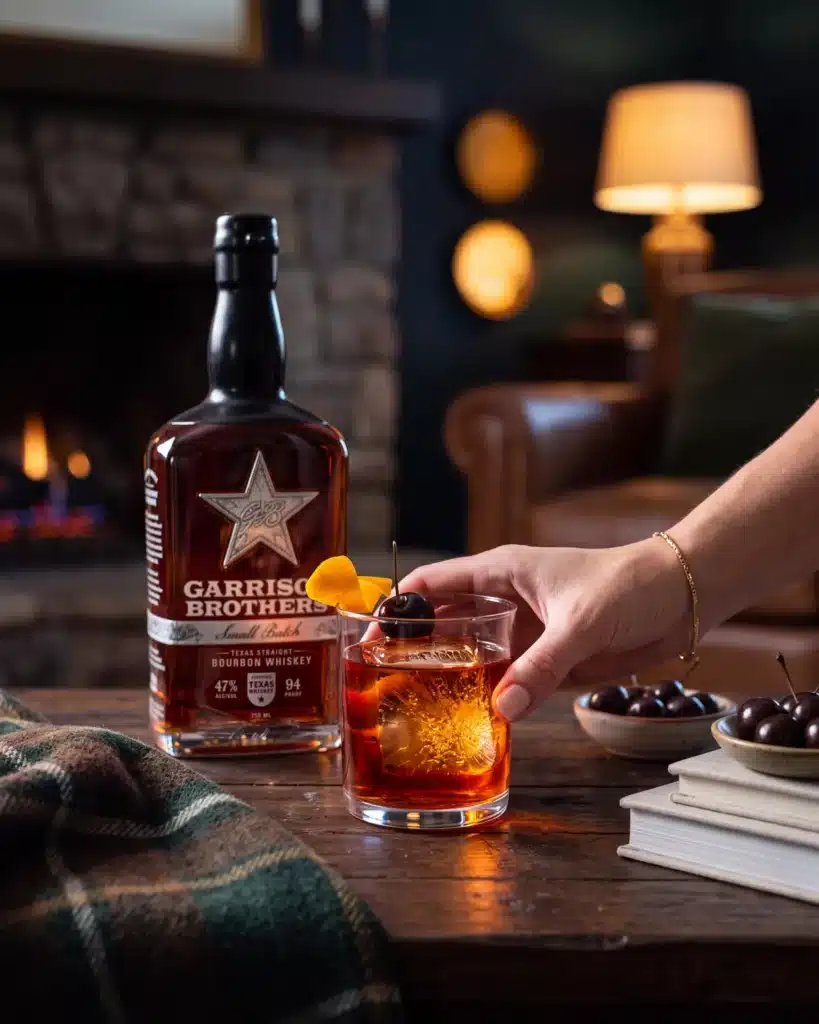 Down To Hearth bourbon cocktail by a stone fireplace with a bottle of Garrison Brothers Small Batch Bourbon
