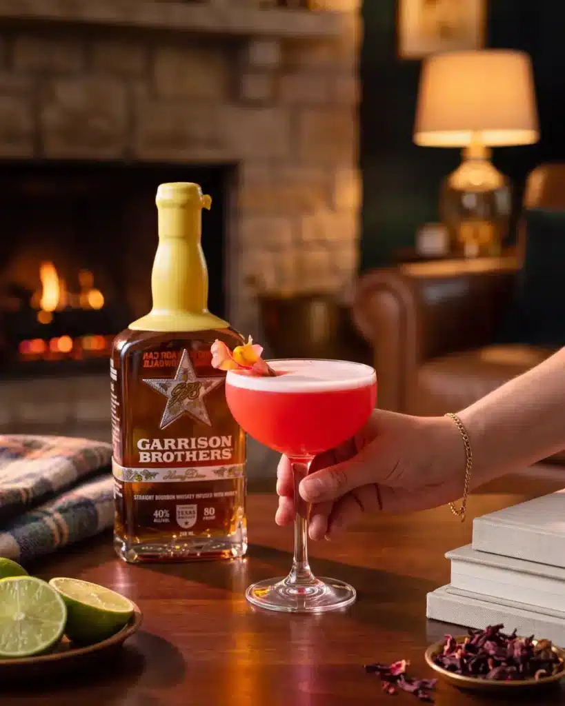Honey Hye-Biscus bourbon cocktail by a stone fireplace with a bottle of Garrison Brothers HoneyDew Bourbon