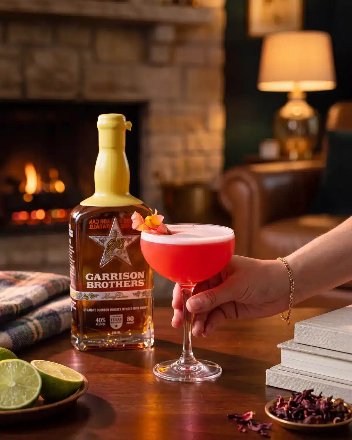 Honey Hye-Biscus bourbon cocktail by a stone fireplace with a bottle of Garrison Brothers HoneyDew Bourbon
