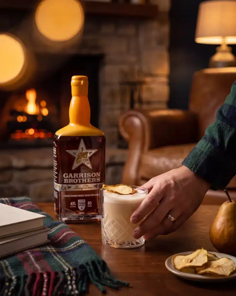 Pearis, Texas bourbon cocktail by a stone fireplace with a bottle of Garrison Brothers HoneyDew Bourbon